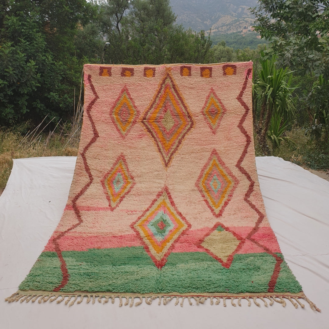 Person holding up a pink and green Moroccan rug outdoors, displaying its 194x293 cm size - a bold blush pink field occupying the upper two-thirds transitioning into a solid jade green band across the bottom, with large concentric diamond medallions in yellow, orange and mint stacked vertically, and a brown zigzag line running down both side borders