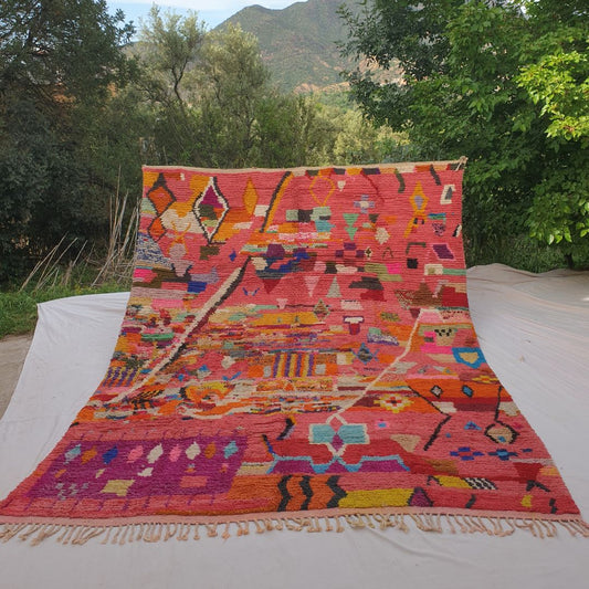 Person holding up a large coral pink Boujad Moroccan rug outdoors between trees, displaying its 293×386 cm size — a warm salmon-coral background densely covered with multicolor geometric motifs in orange, yellow, teal, purple, blue, green and black, an explosion of Berber artistry