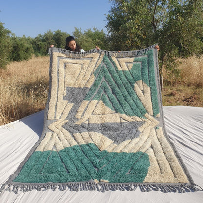 Person holding up a luxurious Beni Mrirt Moroccan rug outdoors, showing its 203×294 cm size with a striking architectural composition — bold teal emerald green concentric diamond and rectangle frames on a cream ivory wool background with soft grey lavender triangular accents, the thick plush Beni Mrirt pile clearly visible