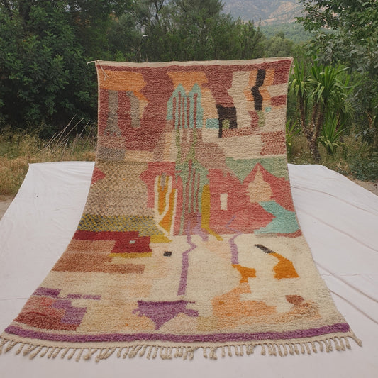 Person holding up a neutral pastel Moroccan rug outdoors, displaying its 195x321 cm size - a predominantly ivory ground with a soft, muted abstract patchwork of dusty mauve, sage, warm tan and ochre colour blocks, delicate purple vertical drip-like line accents, and a solid purple border stripe