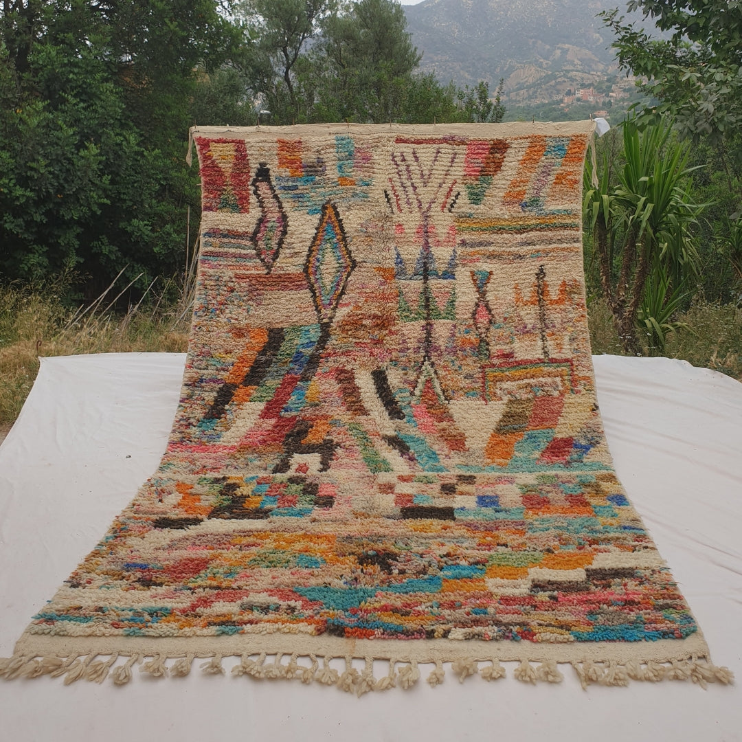 Person holding up a colorful ivory Moroccan rug outdoors, displaying its 187x307 cm size - a dense ivory field covered in multicolor confetti-like flecks of turquoise, orange, pink and red, with a black-outlined diamond motif, a zigzag chevron pattern and a bold checkerboard band running through the middle