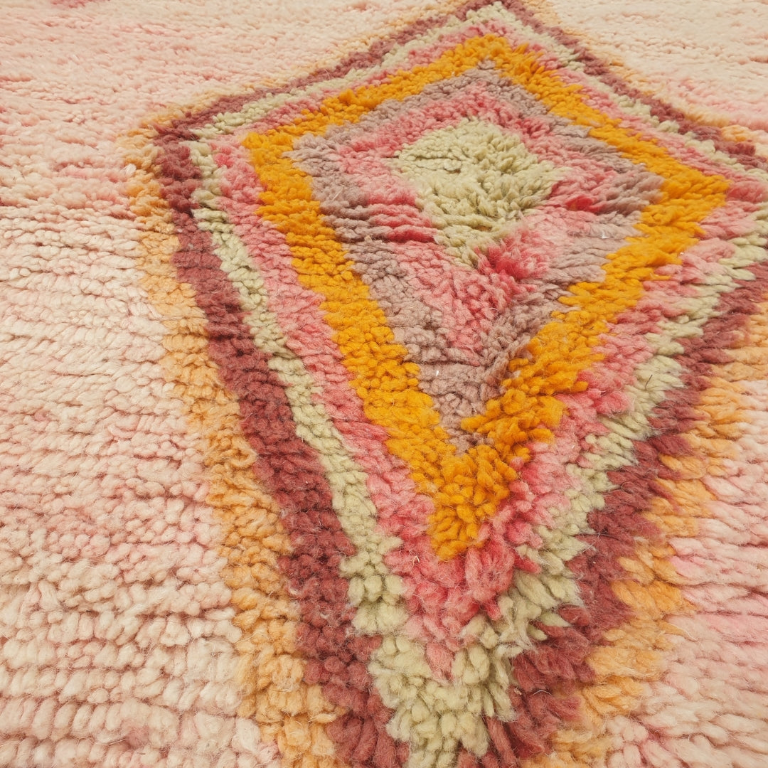 Close-up of a Kasma Moroccan rug showing the soft wool pile with a large concentric diamond medallion - yellow, orange and mint rings nesting inward against the blush pink field, handwoven in 100% wool
