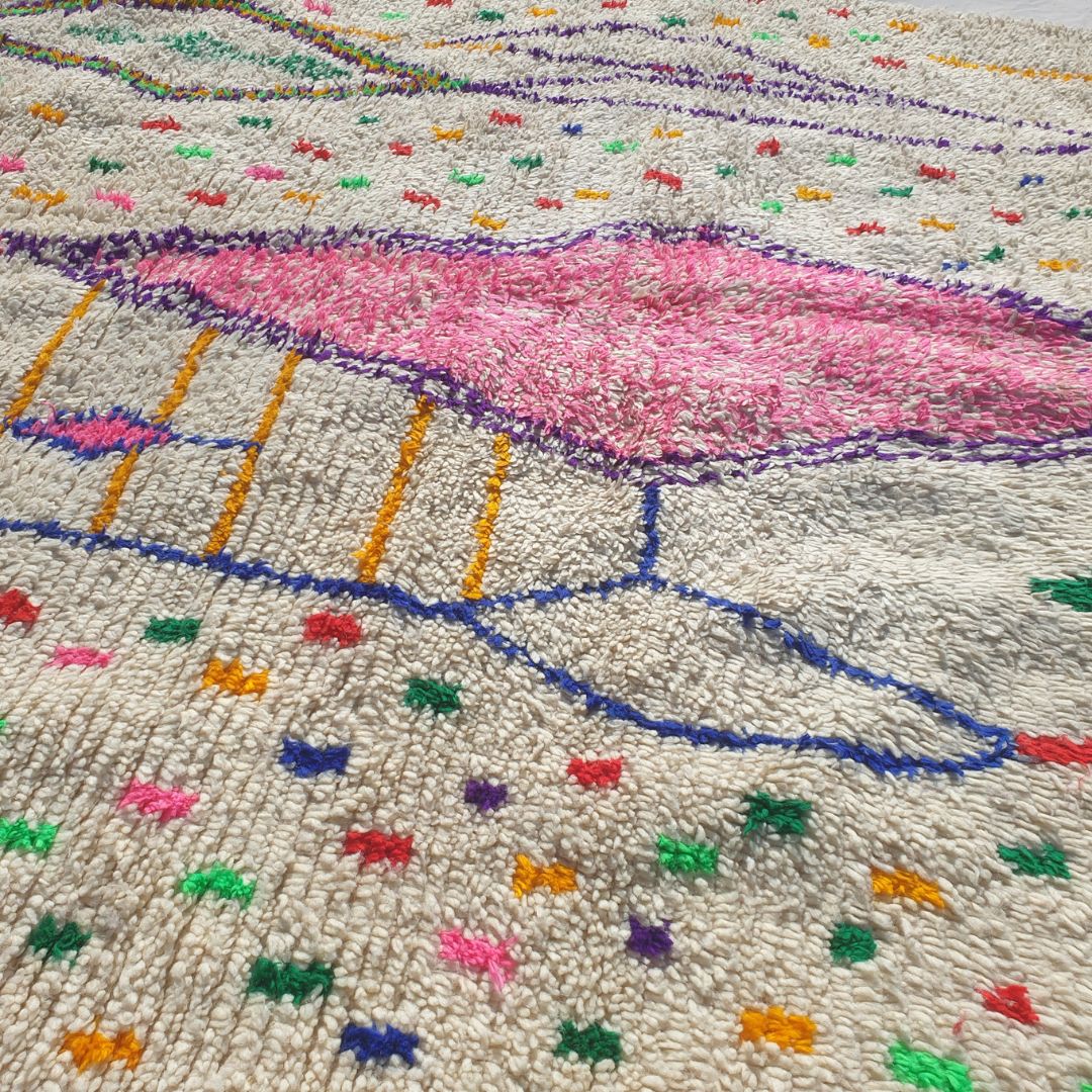 Close-up of a white Azilal Berber rug showing the natural cream wool pile densely scattered with tiny multicolor confetti dots and marks in red, green, orange, yellow and blue — the central rose-pink diamond motif and purple outlined elongated diamonds visible, all handwoven with extraordinary detail and Amazigh symbolism