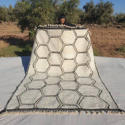 Classic honeycomb geometric 6x9 Beni Ourain Moroccan rug with interconnected hexagons in deep black on soft creamy ivory wool, ultra-fluffy high-pile texture, displayed outdoors in olive grove under blue sky