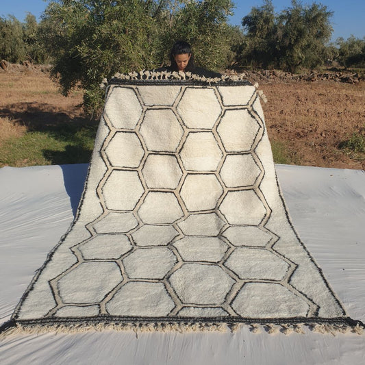 Classic honeycomb geometric 6x9 Beni Ourain Moroccan rug with interconnected hexagons in deep black on soft creamy ivory wool, ultra-fluffy high-pile texture, displayed outdoors in olive grove under blue sky