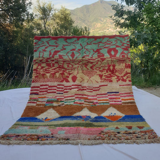 large multicolour Boujad Moroccan rug held up in nature, showing its bold red, teal, brown, blue and pastel abstract landscape bands.