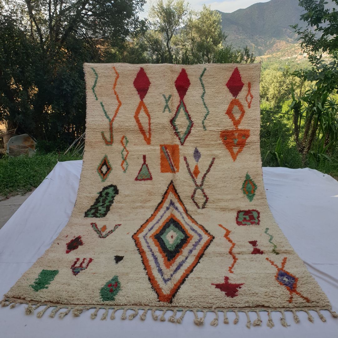 large ivory and colourful Boujad Moroccan rug held up in nature, showing scattered tribal diamond symbols in orange, green, red and purple on a cream ground.
