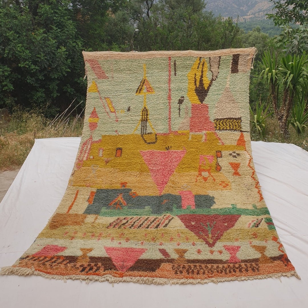 Person holding up a green and mustard Moroccan rug outdoors, displaying its 193x308 cm size - a soft mint sage field transitioning into mustard gold and ochre bands, with a dense pictorial composition of pink inverted triangles, small vessel forms, a bird-like figure and a checkerboard motif
