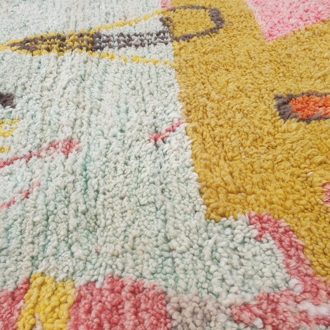 Close-up of a Faycha Moroccan rug showing the soft wool pile with the pictorial narrative composition - pink triangle forms, olive checkerboard blocks and a teal-grey panel visible against the mustard gold field, handwoven in 100% wool