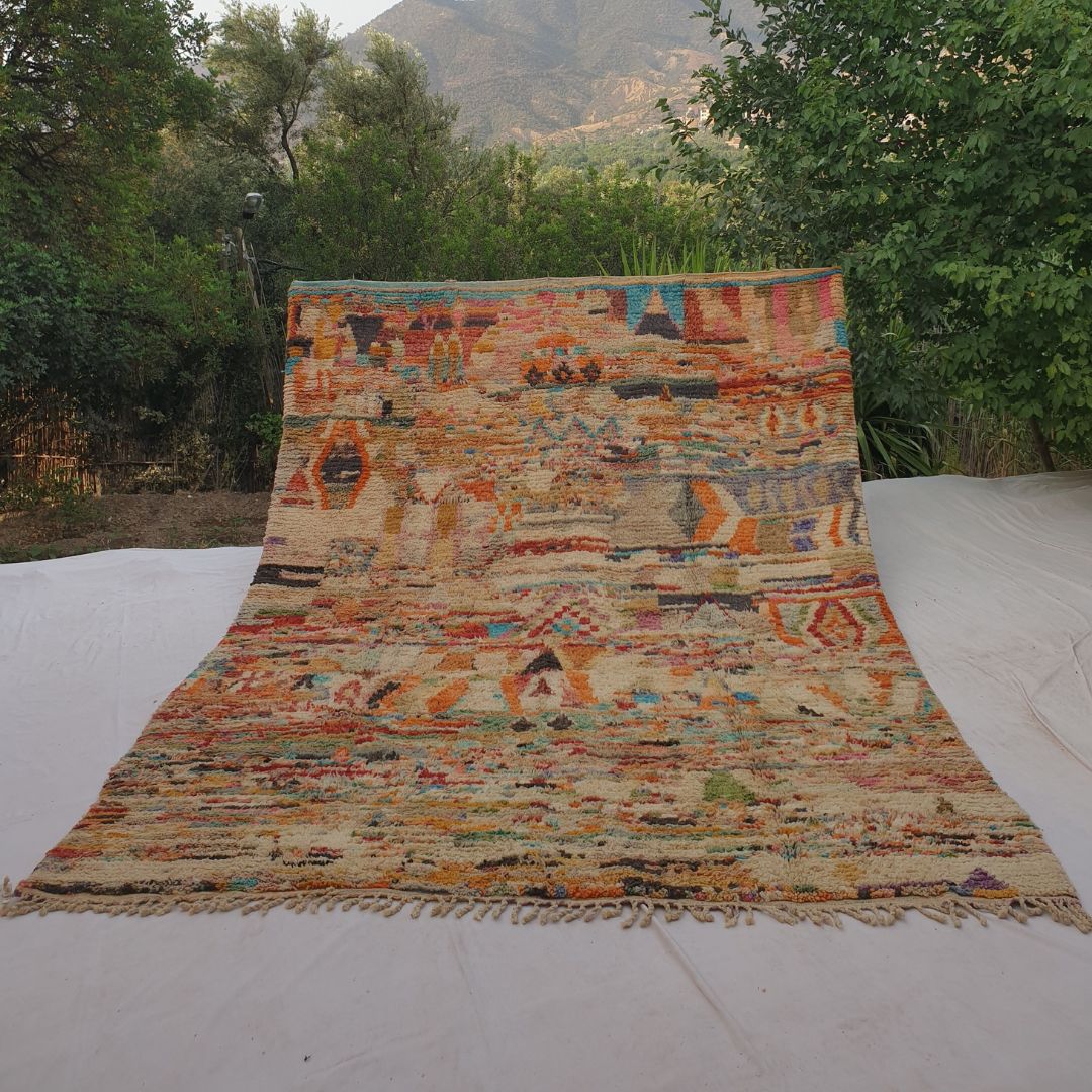 Large 9x12 Boujad Moroccan rug displaying vibrant abstract tribal motifs in warm reds, oranges, pinks, yellows, blues, greens and black on creamy ivory wool, high-pile texture, laid out outdoors with mountain view