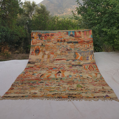 Large 9x12 Boujad Moroccan rug displaying vibrant abstract tribal motifs in warm reds, oranges, pinks, yellows, blues, greens and black on creamy ivory wool, high-pile texture, laid out outdoors with mountain view