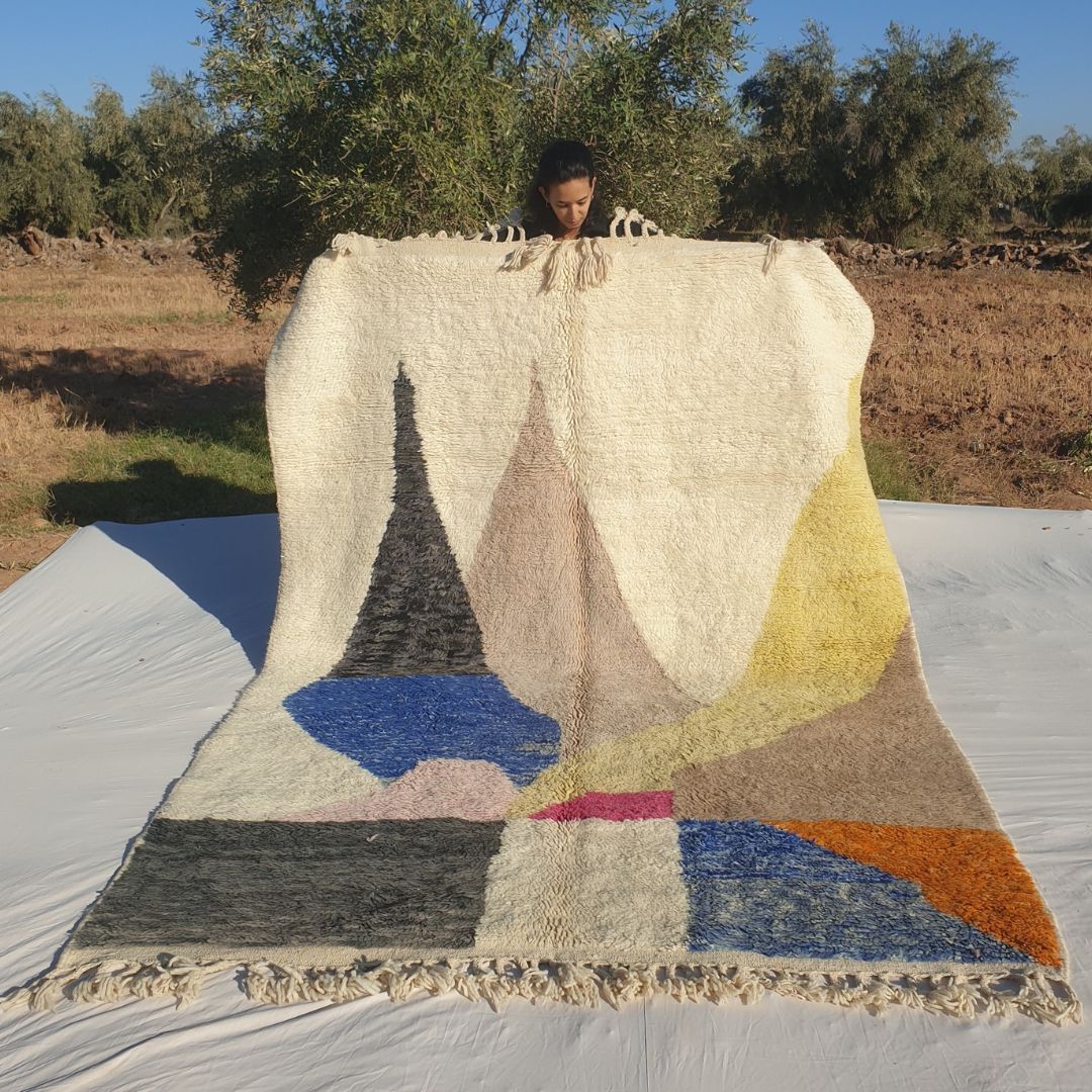 Stunning 6x9 abstract Beni Ourain Moroccan rug with bold colorful geometric shapes in blue, gray, pink, orange and beige on creamy ivory wool background, high-pile fluffy texture, held outdoors in olive grove