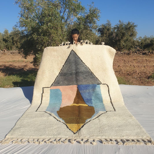 Striking 6x9 abstract Beni Ourain Moroccan rug featuring large geometric diamond and angular shapes in gray, blue, pink, beige, brown and yellow on creamy ivory wool, ultra-fluffy high-pile texture, displayed outdoors in olive grove under blue sky