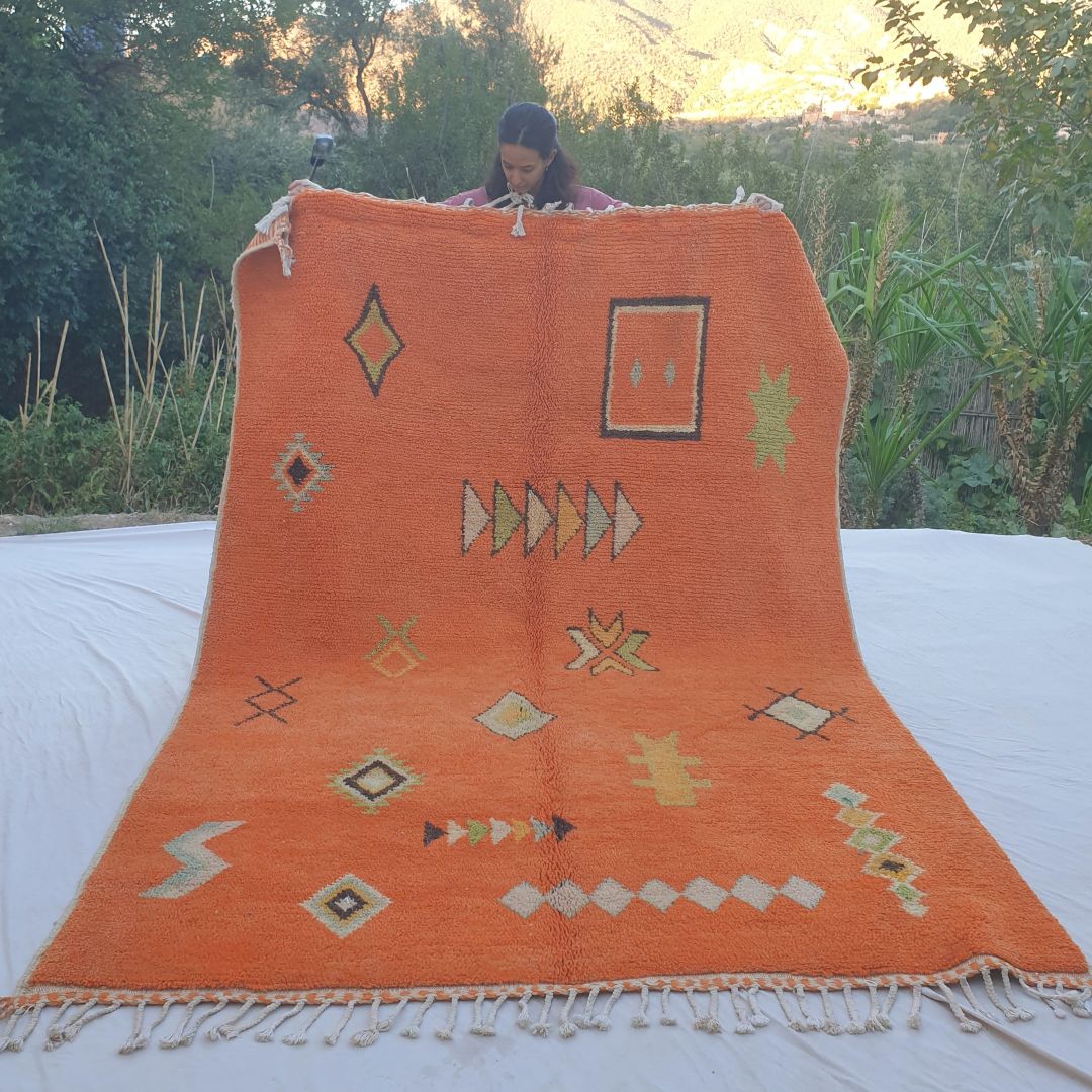 Vibrant orange 6x9 Boujad Moroccan rug featuring bold tribal geometric patterns including diamonds, arrows, crosses and abstract motifs in black, green, yellow and cream, held outdoors against mountain backdrop