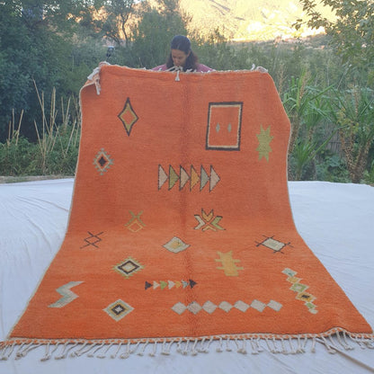 Vibrant orange 6x9 Boujad Moroccan rug featuring bold tribal geometric patterns including diamonds, arrows, crosses and abstract motifs in black, green, yellow and cream, held outdoors against mountain backdrop