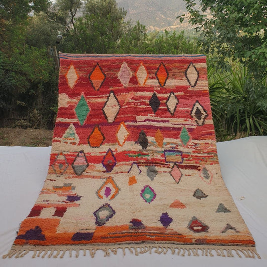 Vibrant red 6x9 Boujad Moroccan rug featuring bold diamond tribal patterns in orange, pink, teal, blue, green and black on creamy ivory wool, high-pile texture, displayed outdoors with mountain backdrop
