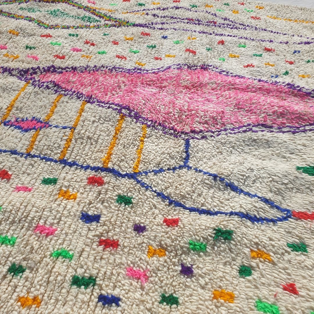 Close-up of a white Azilal Berber rug showing the natural cream wool pile densely scattered with tiny multicolor confetti dots and marks in red, green, orange, yellow and blue — the central rose-pink diamond motif and purple outlined elongated diamonds visible, all handwoven with extraordinary detail and Amazigh symbolism