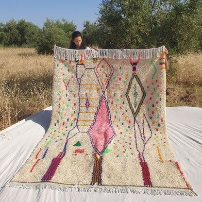 Person holding up a white Azilal Moroccan rug outdoors, showing its 195×282 cm size — a warm cream natural wool base scattered with joyful multicolor confetti dots in red, green, orange, yellow, blue and pink, with a large central rose-pink diamond, elongated purple diamond outlines, green outlined upper diamonds and bold navy and red vertical accent lines at the bottom