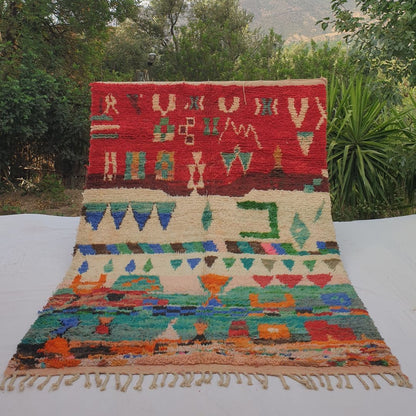 Vibrant 6x9 Boujad Moroccan rug featuring bold tribal symbols, diamonds, arrows, geometric shapes and abstract motifs in rich red, orange, pink, green, blue and black on creamy ivory wool, high-pile texture, displayed outdoors with mountain view