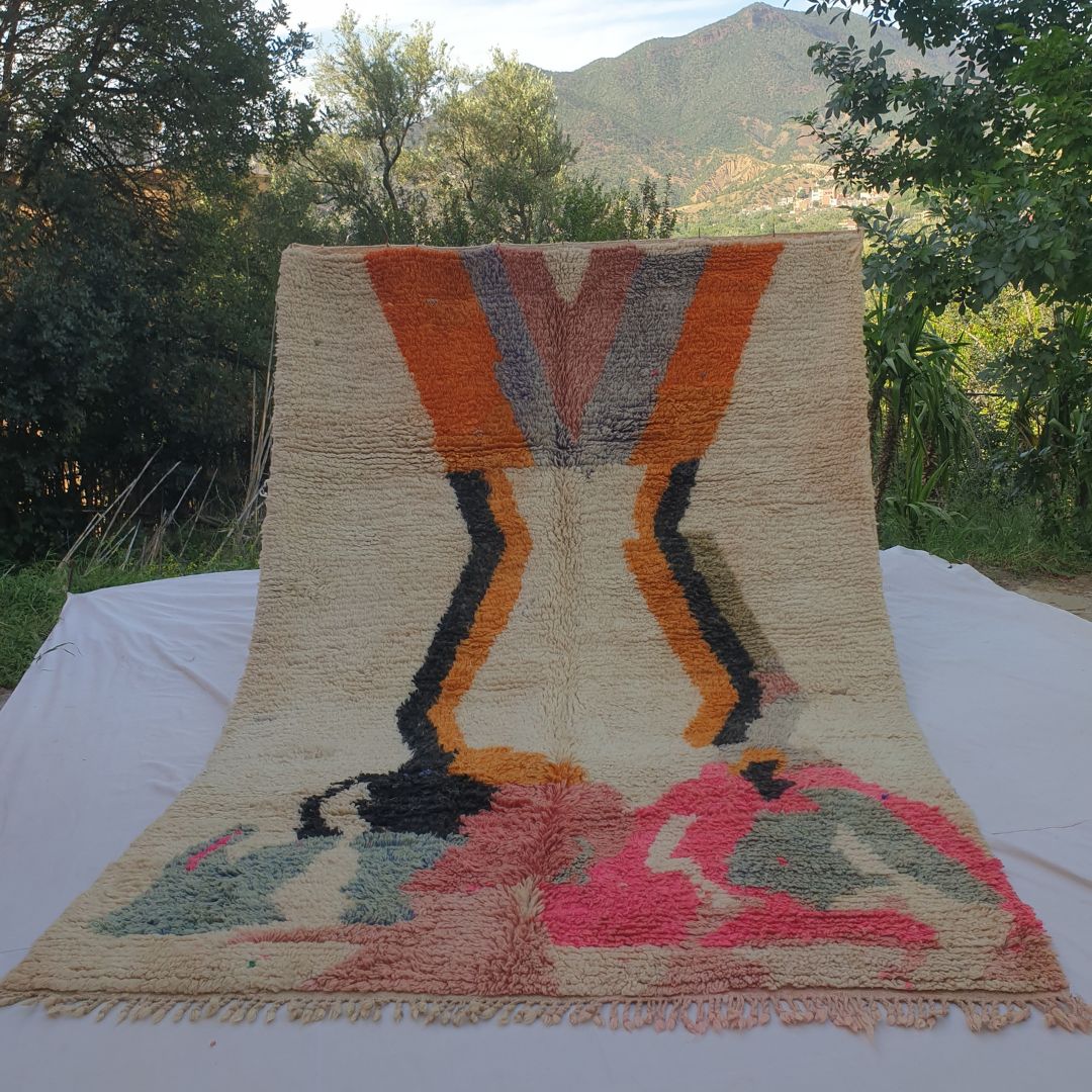 large ivory Boujad Moroccan rug held up in nature, showing two dramatic orange lightning bolt zigzag columns descending from the top with blush-pink and grey triangles, and large abstract hot pink, slate blue and black shapes below.