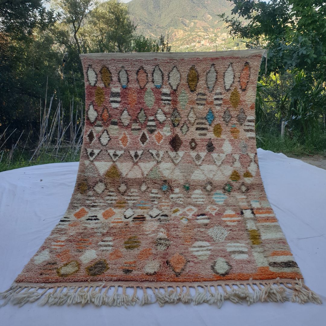 large rose-pink Moroccan Boujaad-style rug held up in nature, showing a densely scattered field of diamond and oval motifs in ivory, mustard gold, teal and orange on a warm pink-terracotta ground.