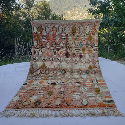 large rose-pink Moroccan Boujaad-style rug held up in nature, showing a densely scattered field of diamond and oval motifs in ivory, mustard gold, teal and orange on a warm pink-terracotta ground.