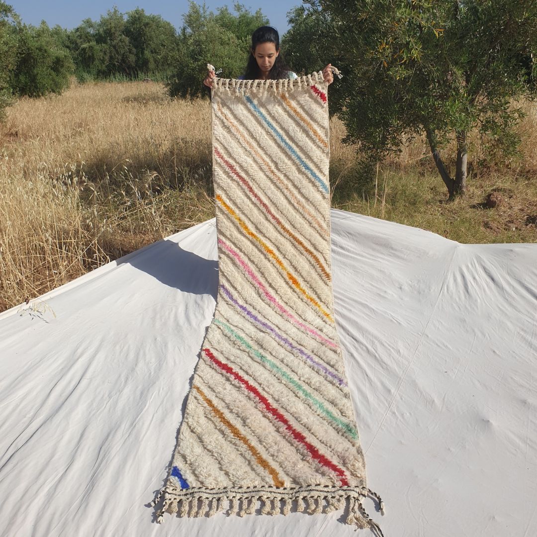 Person holding up a white Beni Ourain Moroccan runner rug outdoors, 73x303 cm - ivory natural wool base covered with bold multicolor diagonal stripes in yellow, orange, pink, purple, teal, red and blue running across the entire width at a consistent angle, creating a dynamic full-field pattern with natural fringe at both ends