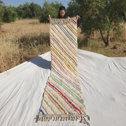 Person holding up a white Beni Ourain Moroccan runner rug outdoors, 73x303 cm - ivory natural wool base covered with bold multicolor diagonal stripes in yellow, orange, pink, purple, teal, red and blue running across the entire width at a consistent angle, creating a dynamic full-field pattern with natural fringe at both ends