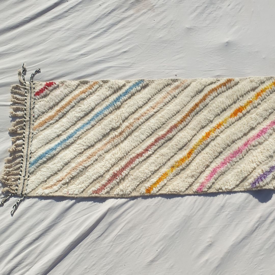 Bottom half of a white Beni Ourain Moroccan runner with bold multicolor diagonal stripes in yellow, orange, pink, purple and teal running from edge to edge across the full ivory wool pile, with natural knotted fringe
