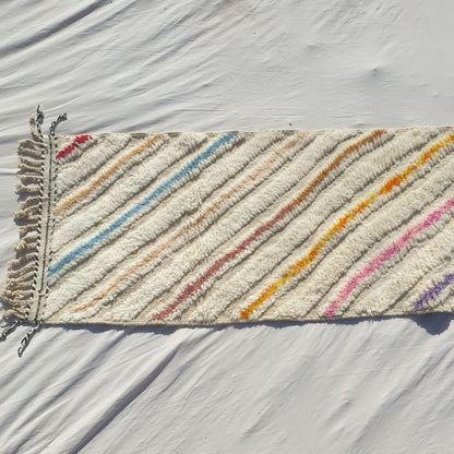 Bottom half of a white Beni Ourain Moroccan runner with bold multicolor diagonal stripes in yellow, orange, pink, purple and teal running from edge to edge across the full ivory wool pile, with natural knotted fringe