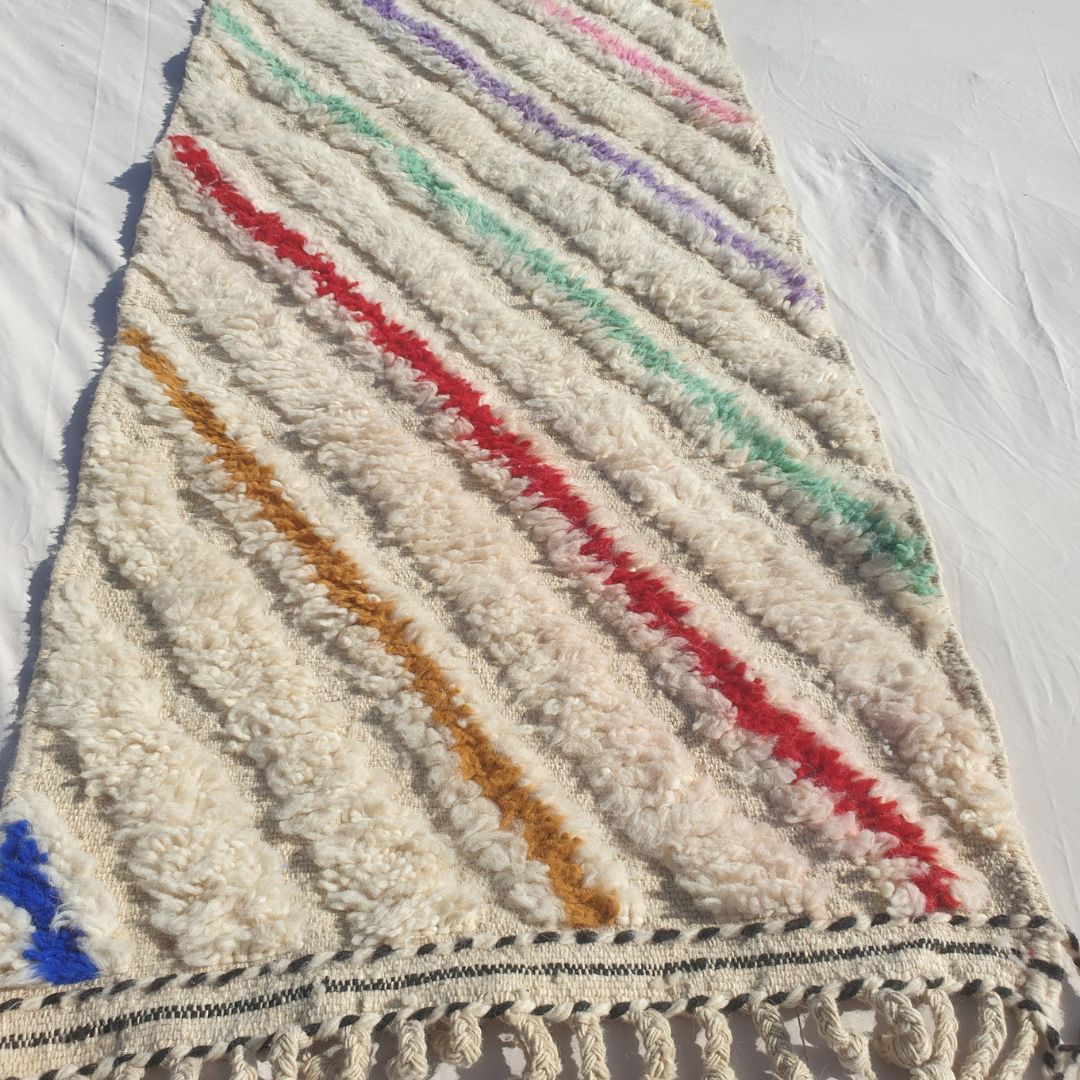 Close-up of a white Beni Ourain runner rug showing the natural ivory wool pile covered with vivid multicolor diagonal stripes - yellow, orange, pink, purple, teal, red and blue bands crossing the field diagonally, each a clean bold stripe, all handwoven in 100% natural Atlas mountain wool