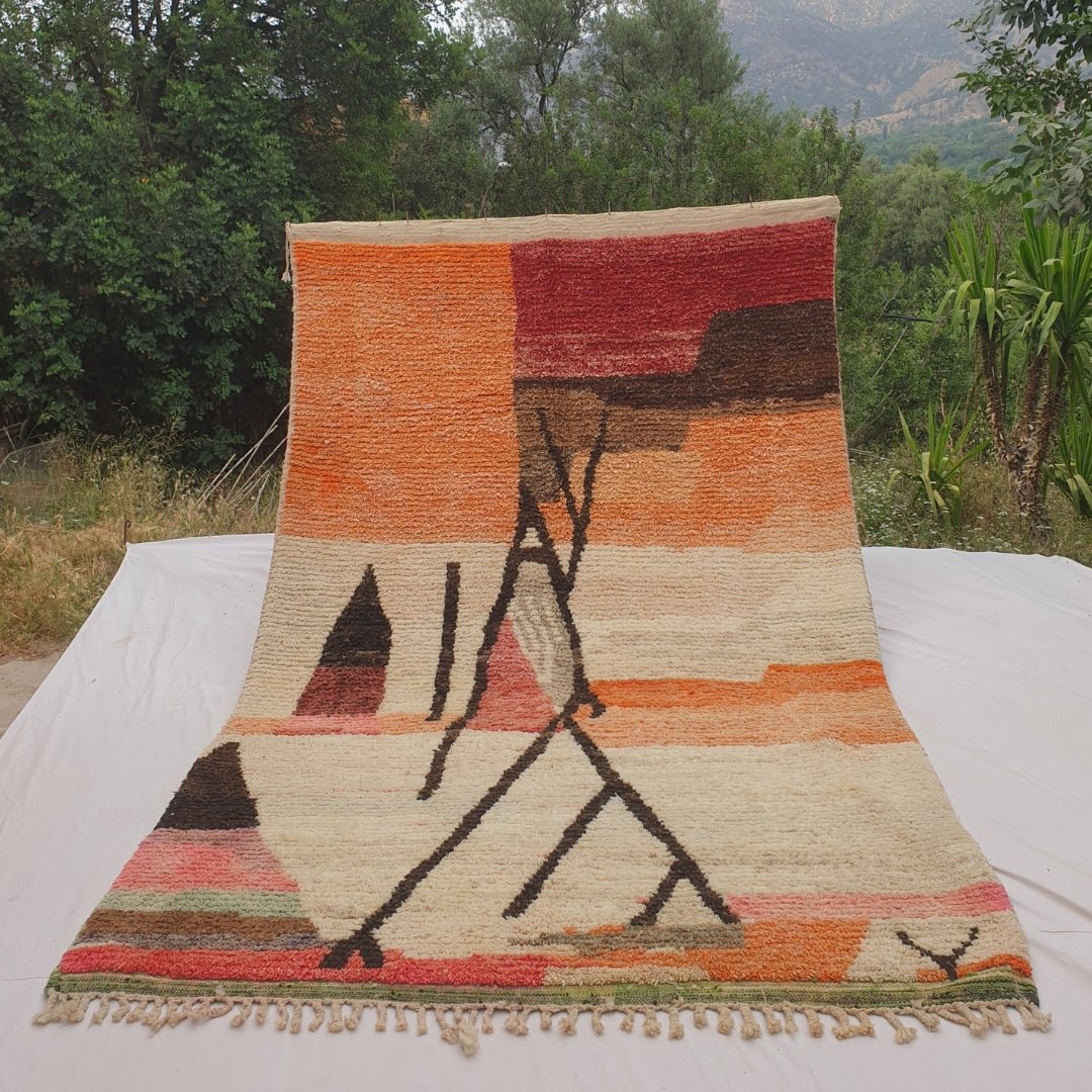 Person holding up an orange and ivory Moroccan rug outdoors, displaying its 197x315 cm size - warm orange and deep red colour fields at the top transitioning to a cream ivory ground, with a bold dark abstract line composition of crossing pole-like forms, a dark triangle, and a multicolour striped band along the lower edge
