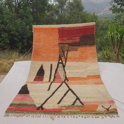 Person holding up an orange and ivory Moroccan rug outdoors, displaying its 197x315 cm size - warm orange and deep red colour fields at the top transitioning to a cream ivory ground, with a bold dark abstract line composition of crossing pole-like forms, a dark triangle, and a multicolour striped band along the lower edge
