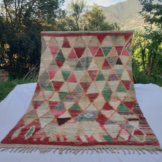 large multicolour harlequin Boujad Moroccan rug held up in nature, showing a bold orange diamond lattice filled with triangles in red, green, ivory, pink, blue and khaki.