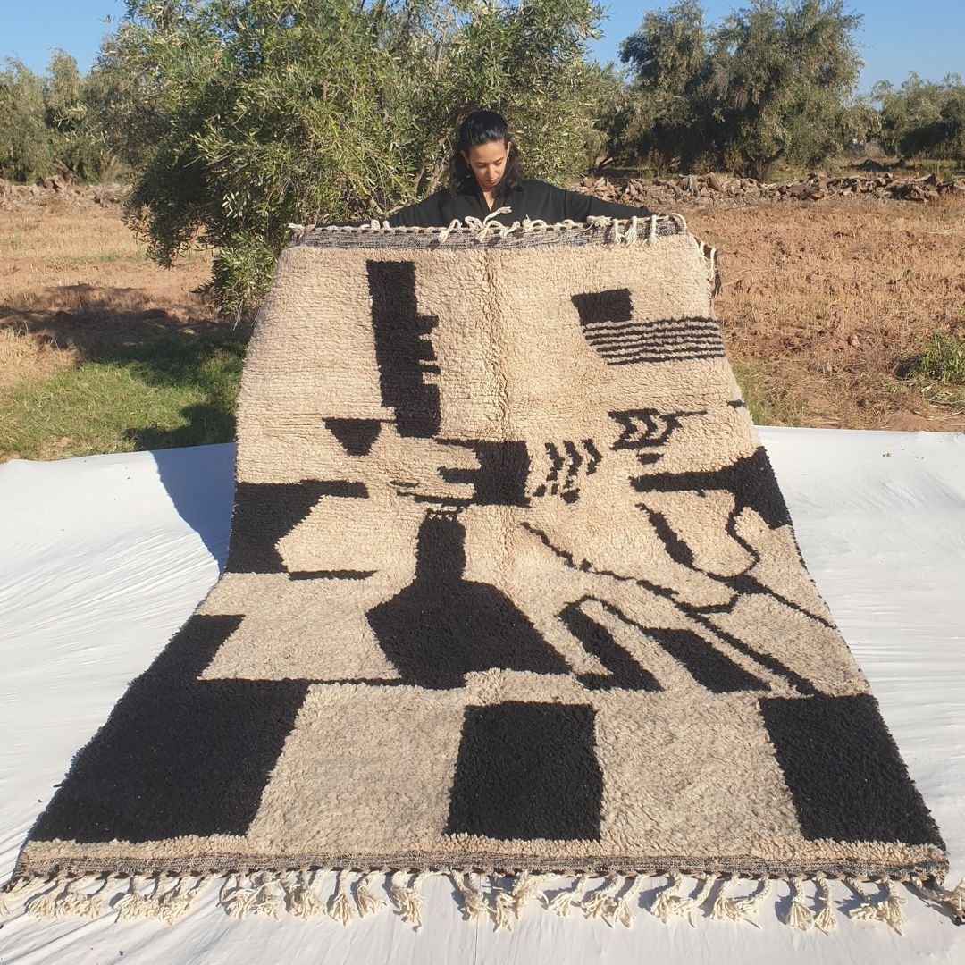 Dramatic 7x9 abstract Beni Ourain Moroccan rug featuring large bold black geometric shapes, tribal forms, and asymmetrical motifs on soft creamy ivory wool, ultra-fluffy high-pile texture, displayed outdoors in olive grove
