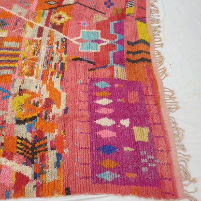 Bottom half of a large coral pink Moroccan wool rug featuring dense handknotted pile with alternating bands of geometric patterns and diamond clusters in vibrant teal, orange, yellow and purple on a warm salmon background, with natural fringe