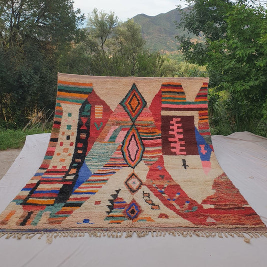Person holding up a large cream and multicolor Boujad Moroccan rug outdoors between trees, displaying its impressive 296×388 cm size with a central column of diamond motifs in teal, pink and orange on an ivory wool background surrounded by vibrant colorful abstract sections
