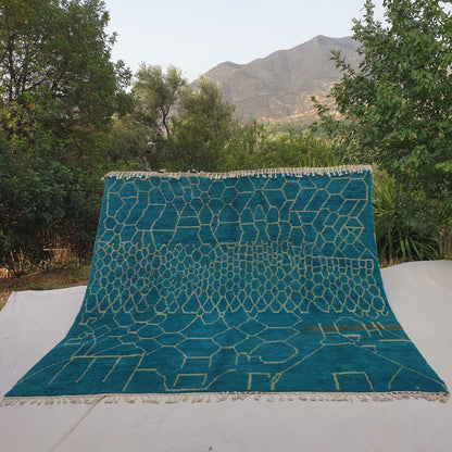 Rare oversized 10x10 square Beni Ourain Moroccan rug featuring intricate honeycomb geometric pattern in rich teal with creamy ivory outlines, ultra-fluffy high-pile texture, laid out outdoors against mountain backdrop and olive trees