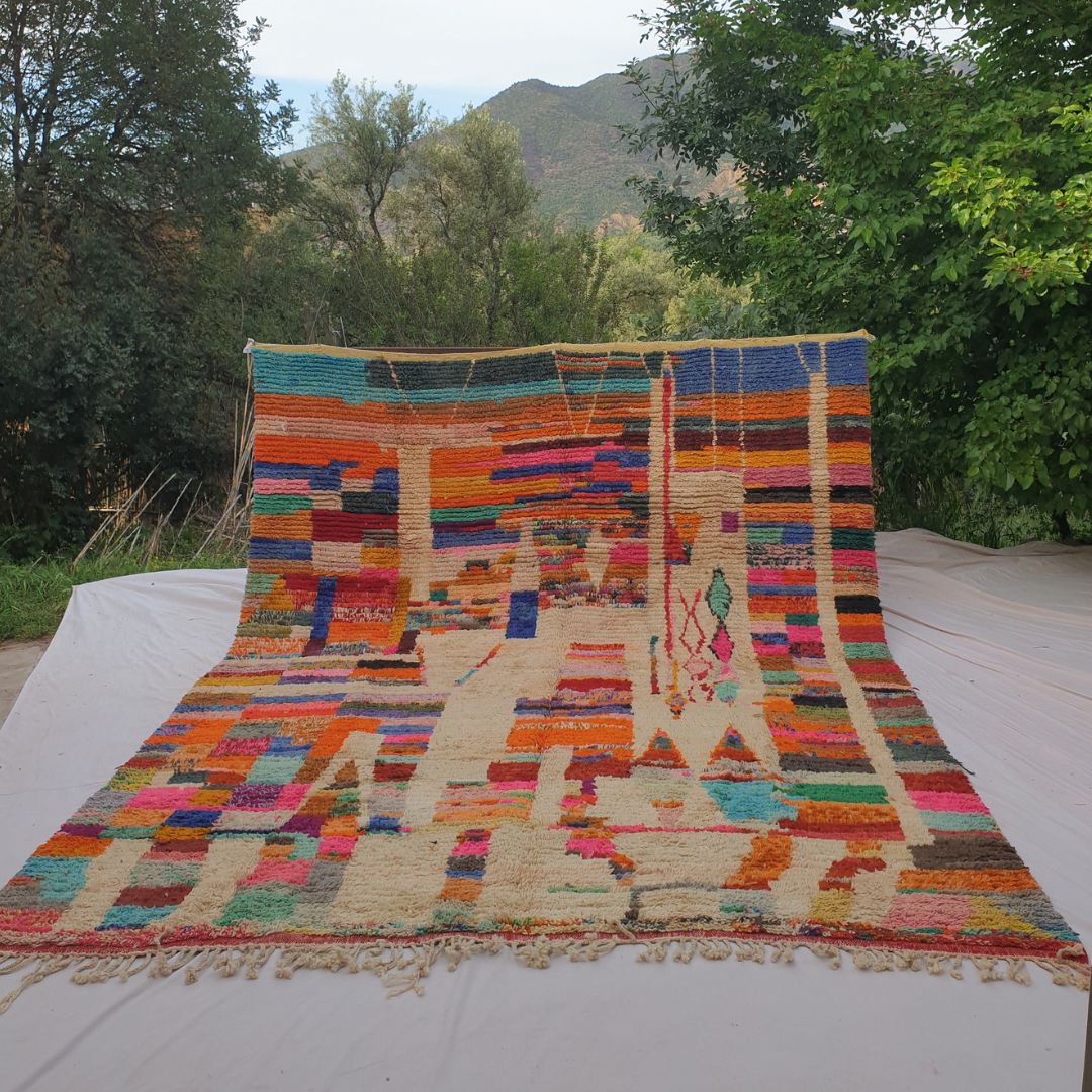 Person holding up a large cream and multicolor Boujad Moroccan rug outdoors between trees, displaying its 311×380 cm size — cream ivory base structured by vertical rivers of natural wool, with dense horizontal stripe bands in orange, teal, red, pink, blue, yellow and green creating a beautiful patchwork mosaic effect