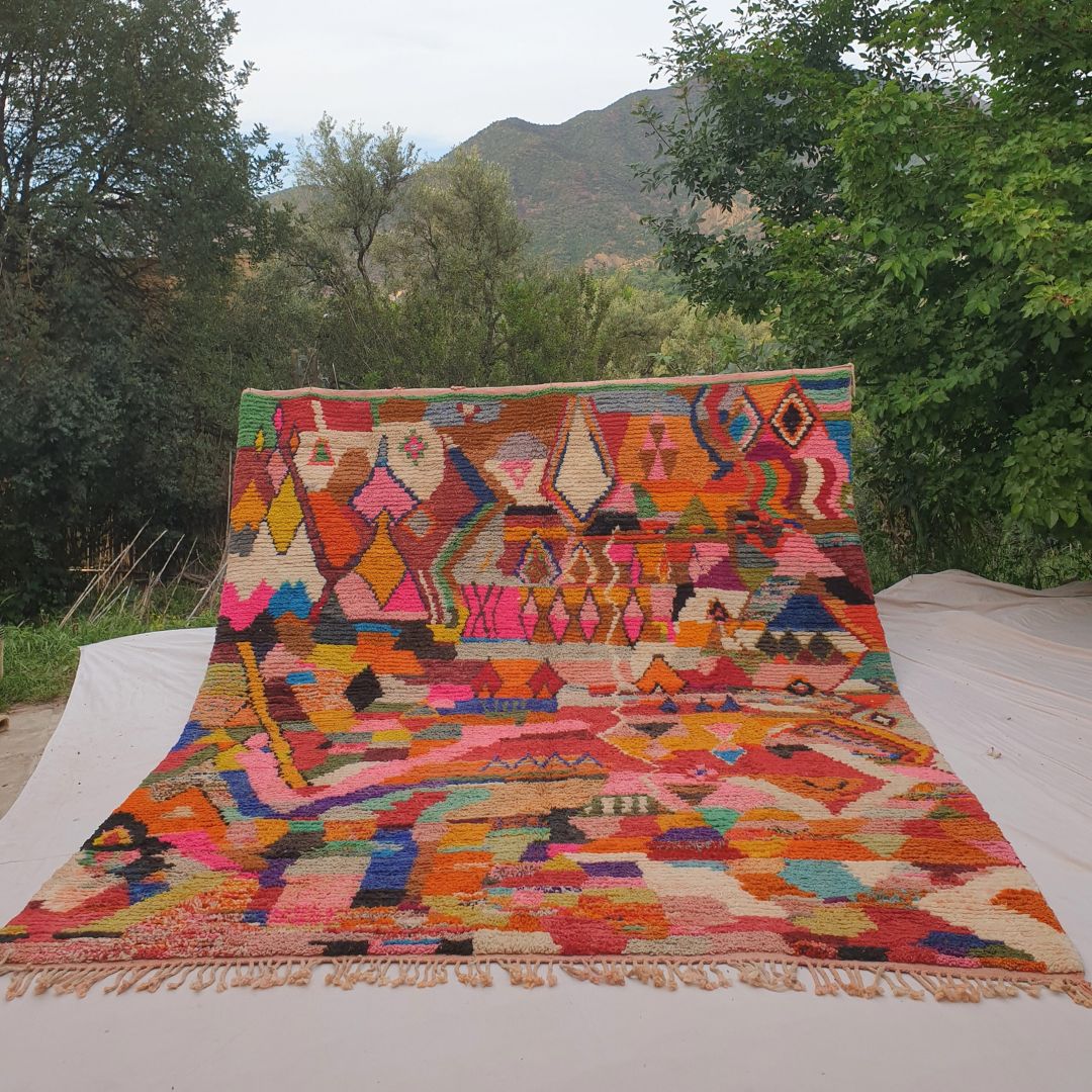 Person holding up an enormous maximalist multicolor Boujad Moroccan rug outdoors between trees, showing its impressive 315×380 cm size — every inch covered in vibrant geometric shapes in orange, pink, red, blue, purple, teal, yellow and green with no dominant background, pure Berber artistic explosion